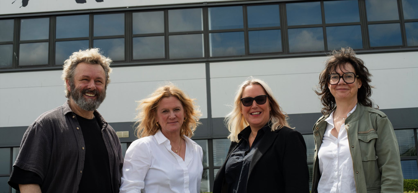 Trivallis Housing Landlord Wales Four adults stand outside a modern building with large windows. Three women and one man, all smiling, wear casual or business-casual clothing. The sky is partly cloudy and reflected in the windows behind them.